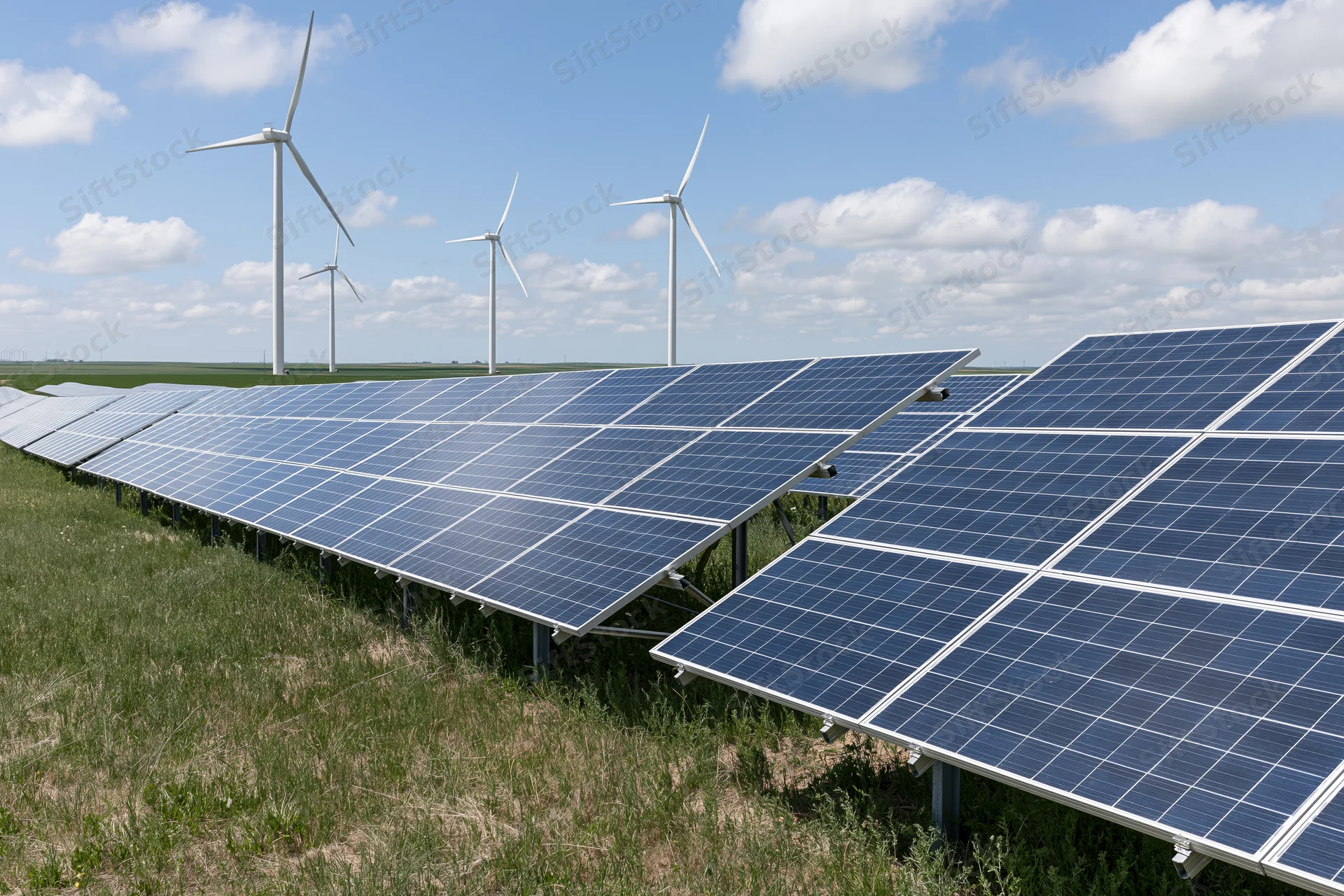 Rows of solar panels angled toward the sky with wind turbines in the background on a grassy field under a blue sky with clouds