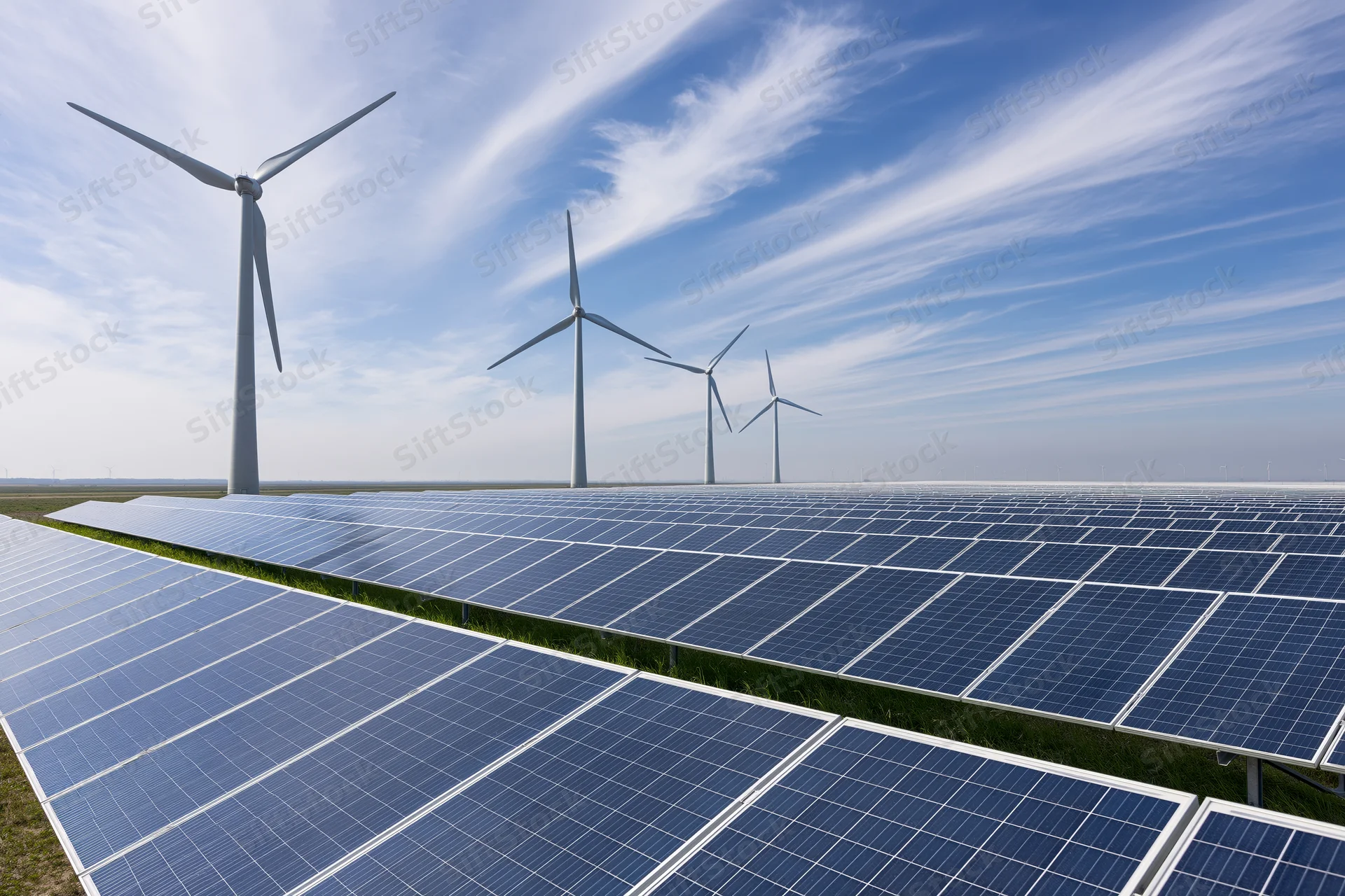Rows of solar panels in a flat rural field with wind turbines in the background under a blue sky with wispy clouds
