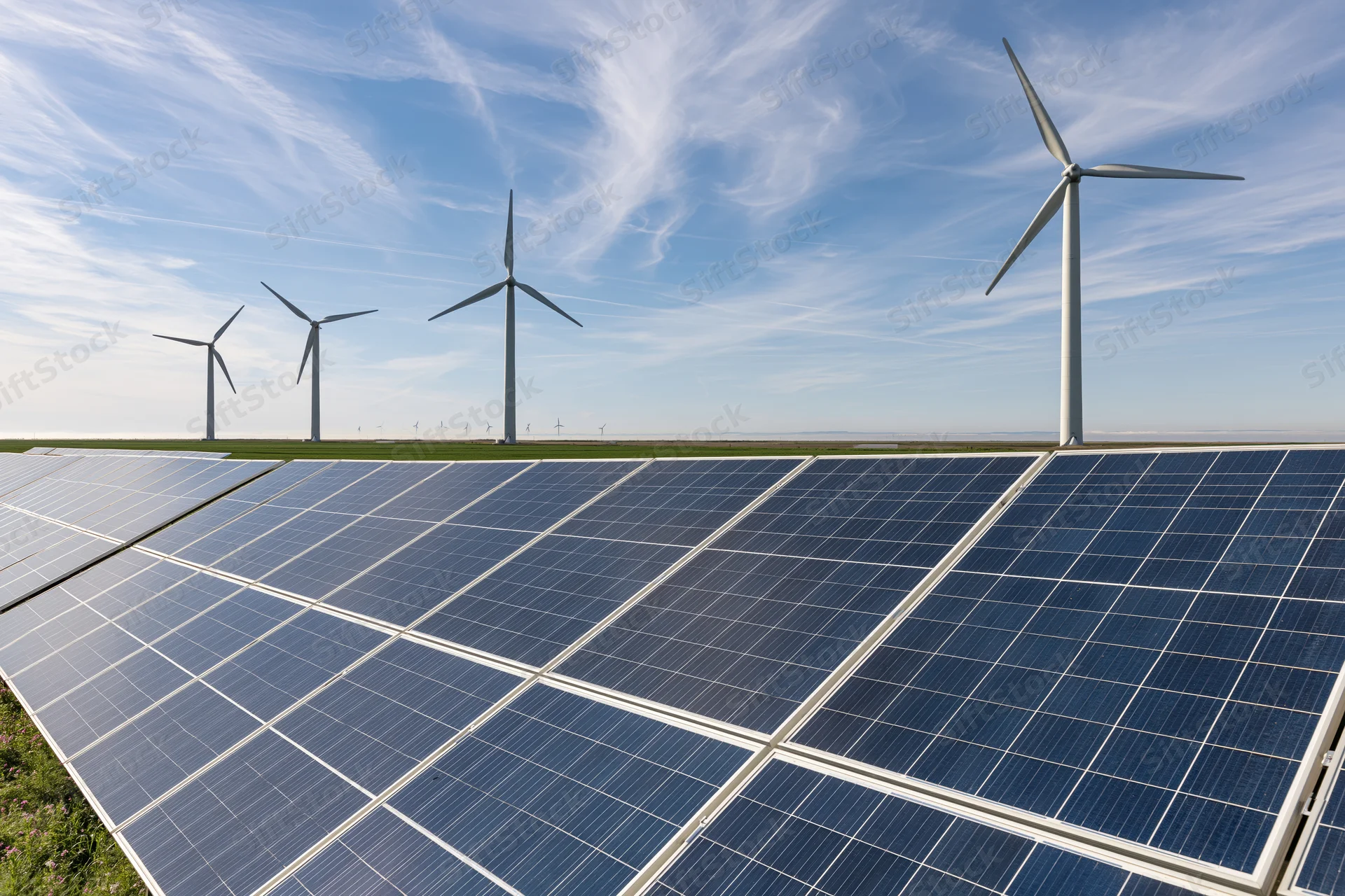 Rows of solar panels in front of wind turbines on a flat rural field under a blue sky with wispy clouds