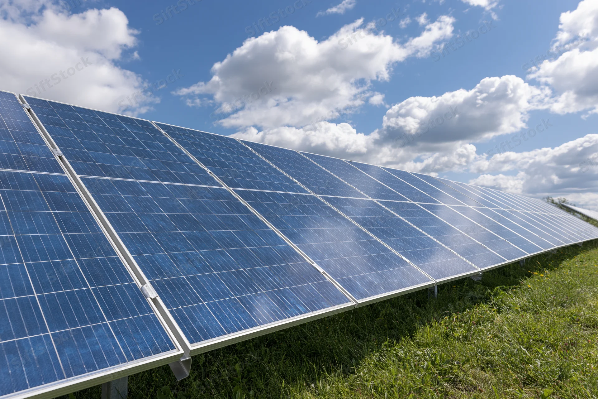 Rows of solar panels installed across a green grass field under a blue sky with white clouds