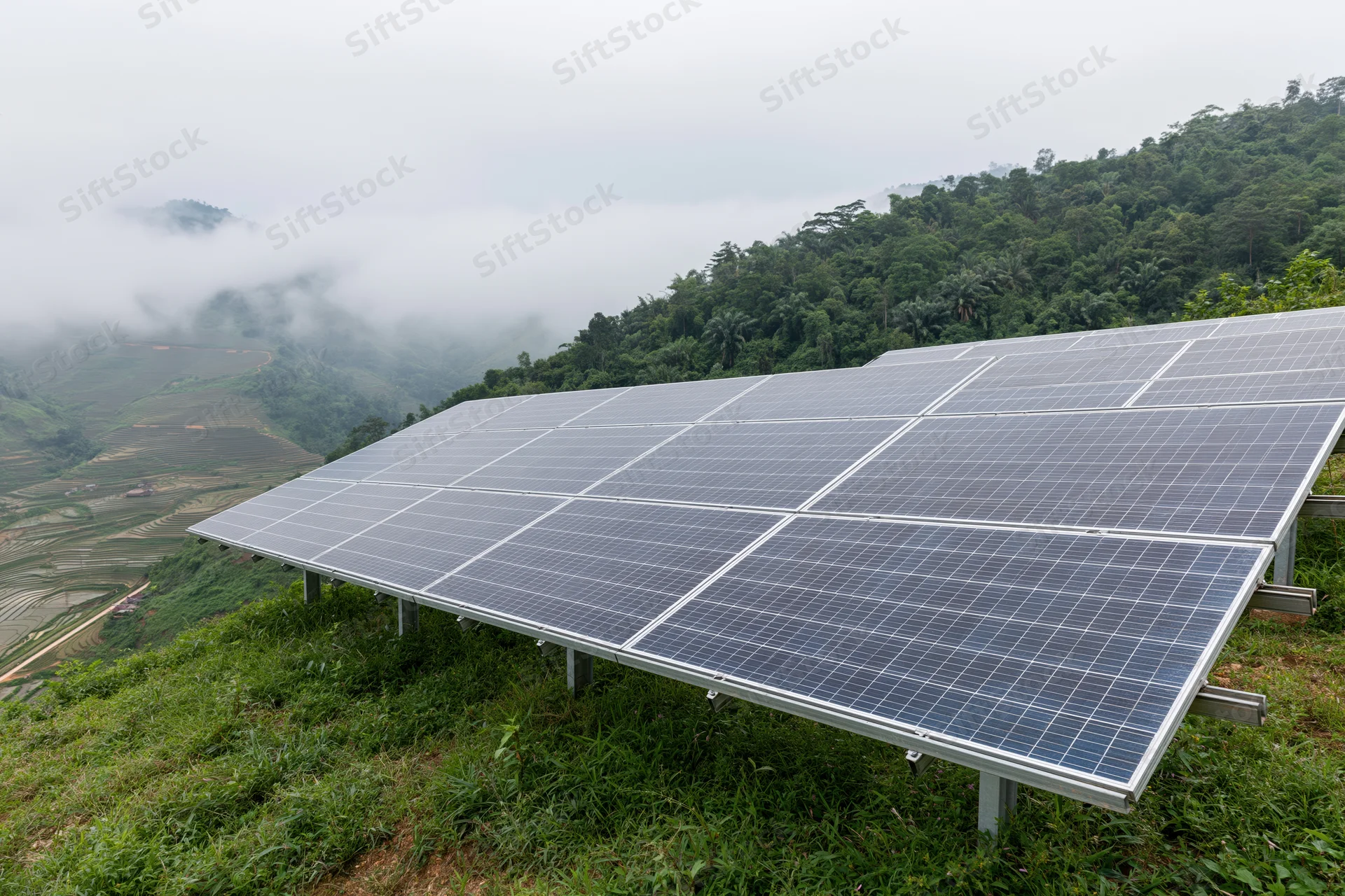 Solar panels installed on a grassy hillside overlooking a misty valley with terraced fields and forested slopes