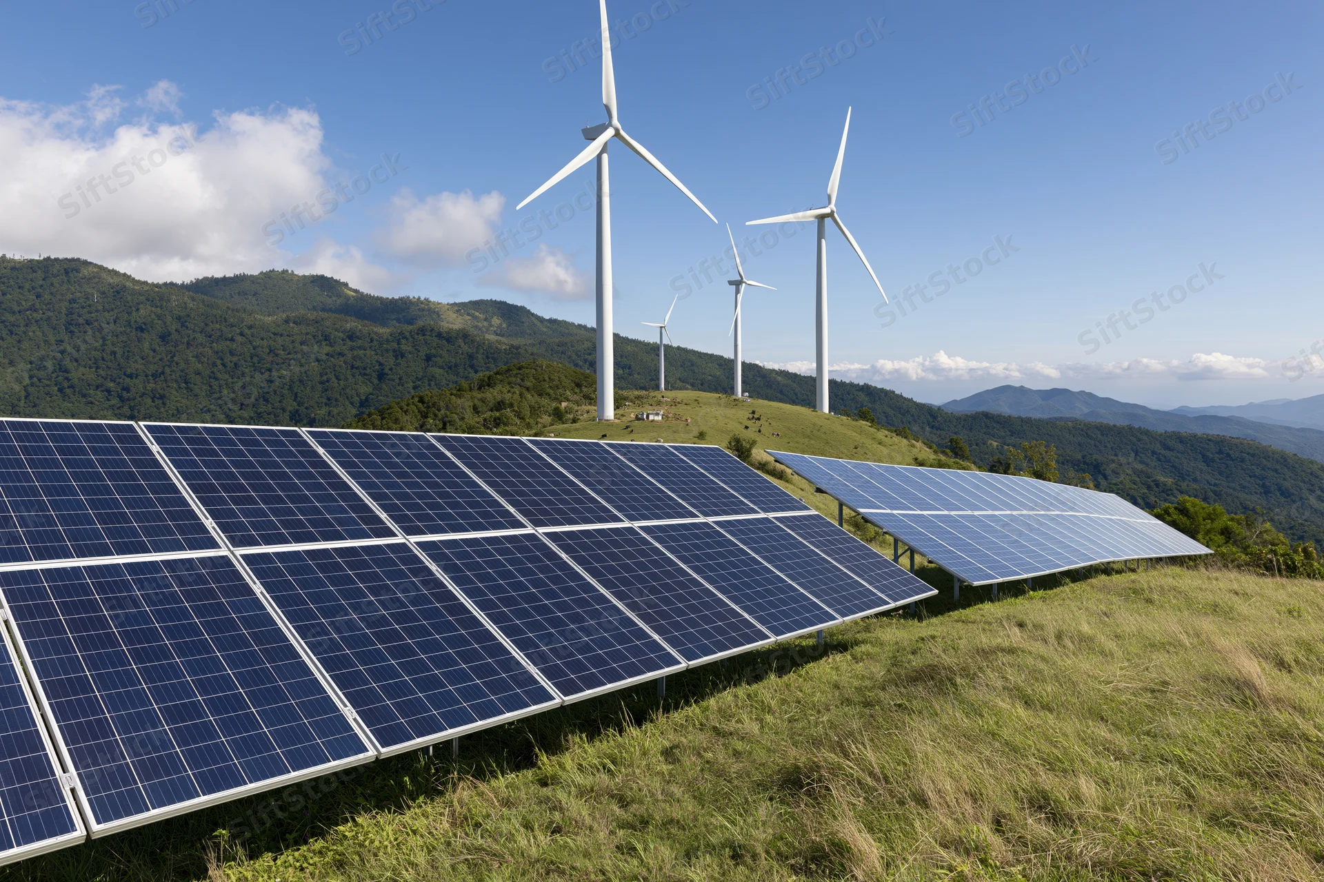 Solar panels and wind turbines arranged on a grassy hillside with mountains in the background under a blue sky with scattered clouds