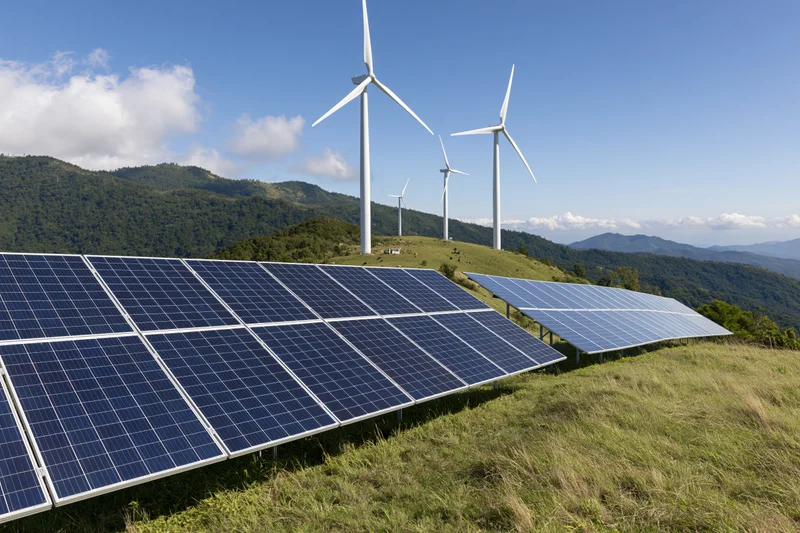 Solar panels and wind turbines arranged on a grassy hillside with mountains in the background under a blue sky with scattered clouds