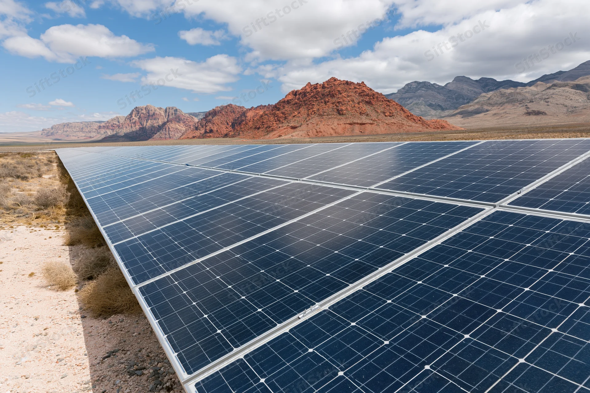 Rows of solar panels arranged in a grid across a flat desert landscape with red rock formations in the background under a partly cloudy sky