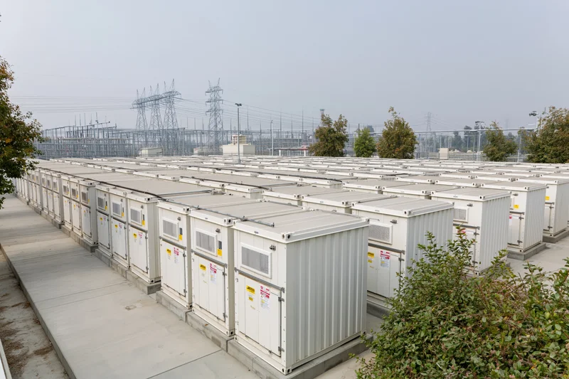 Rows of white industrial battery storage containers arranged in alignment at an outdoor energy facility near a power grid substation