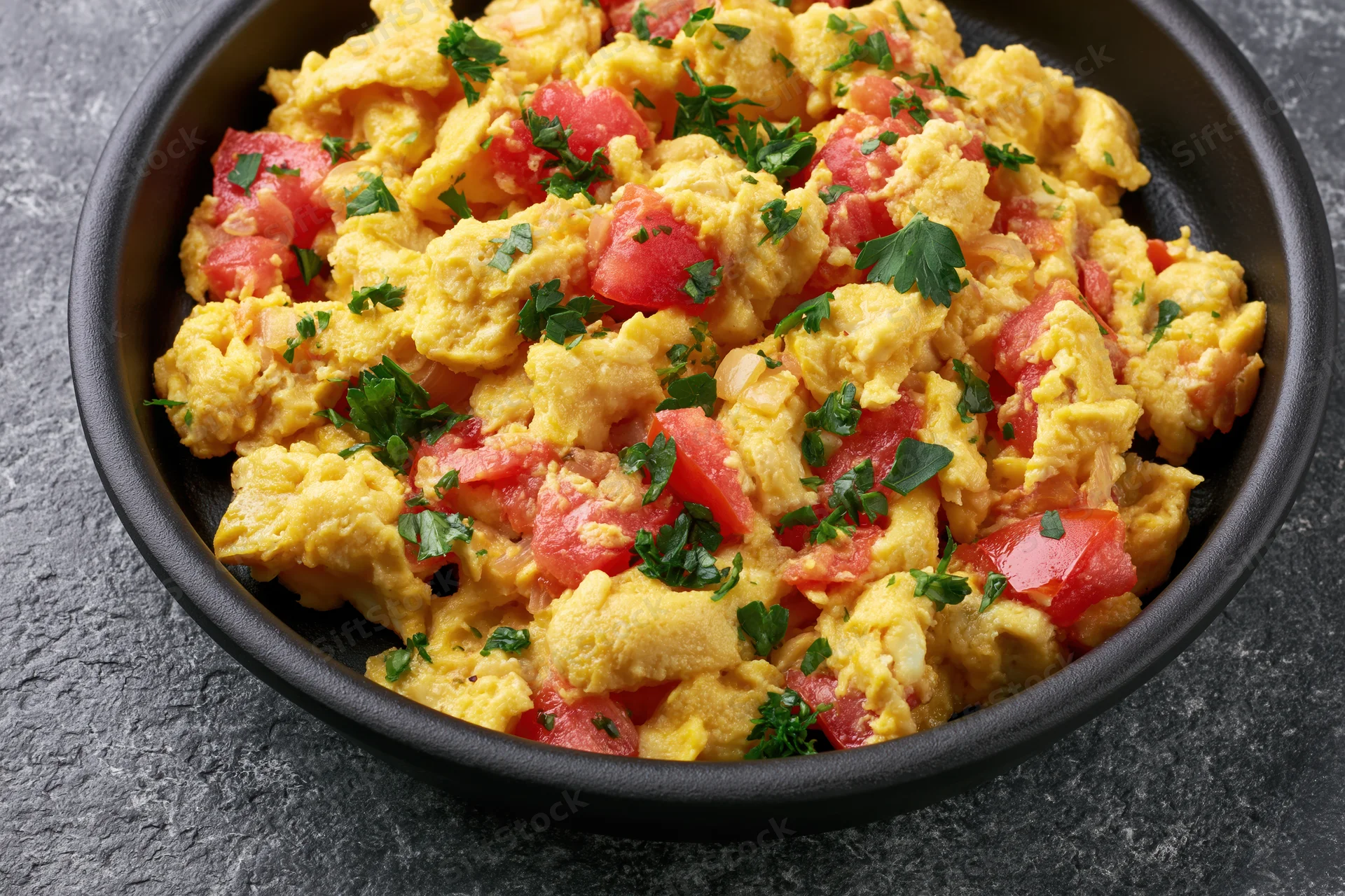 Scrambled eggs with diced tomatoes onions and fresh parsley in a black bowl on a gray surface