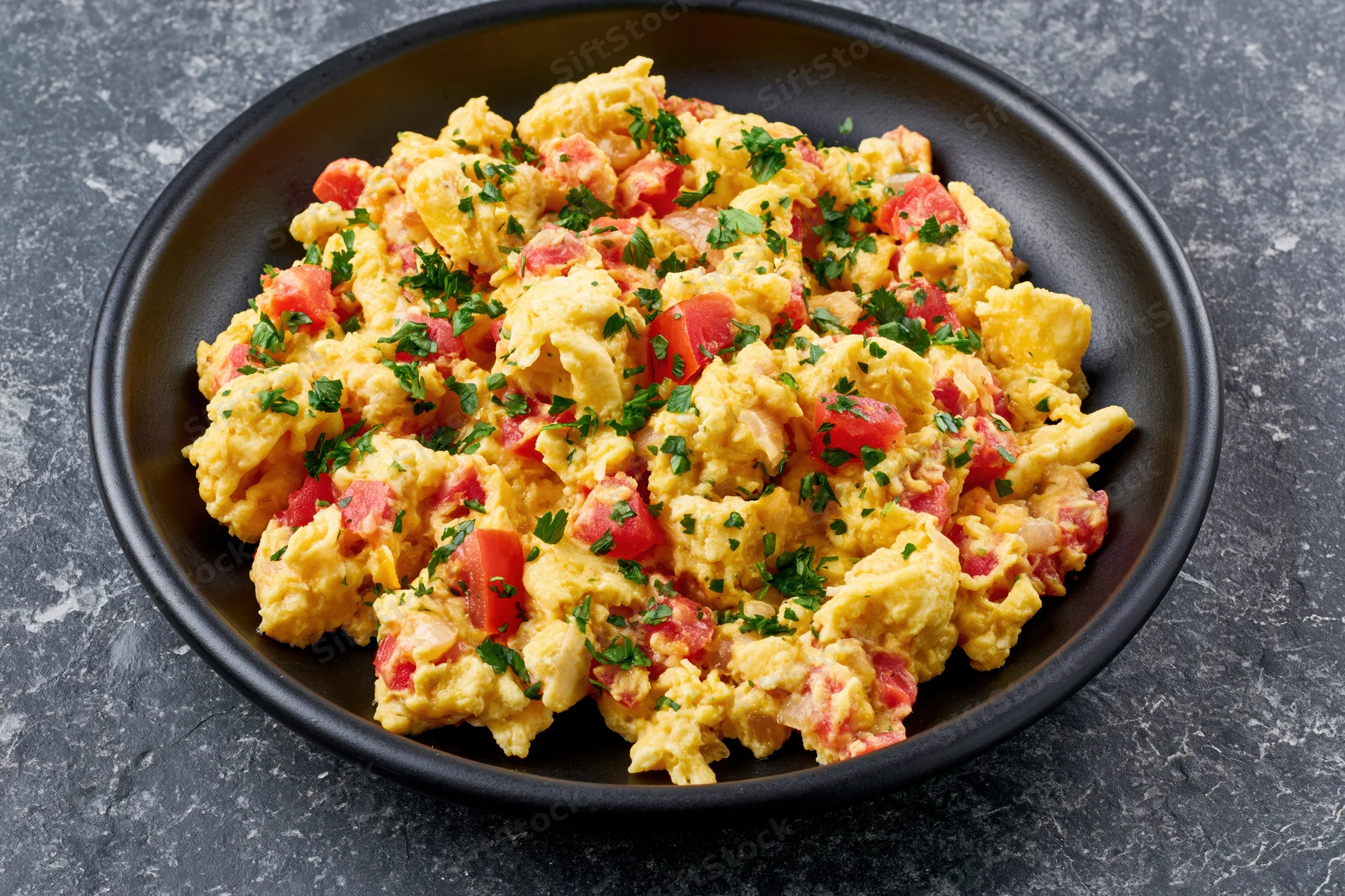 Scrambled eggs with diced tomatoes onions and fresh parsley served in a black bowl on a dark surface