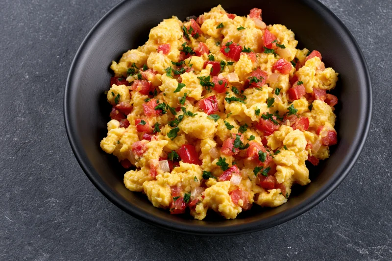 Scrambled eggs with diced tomatoes onions and fresh parsley in a black bowl on dark surface