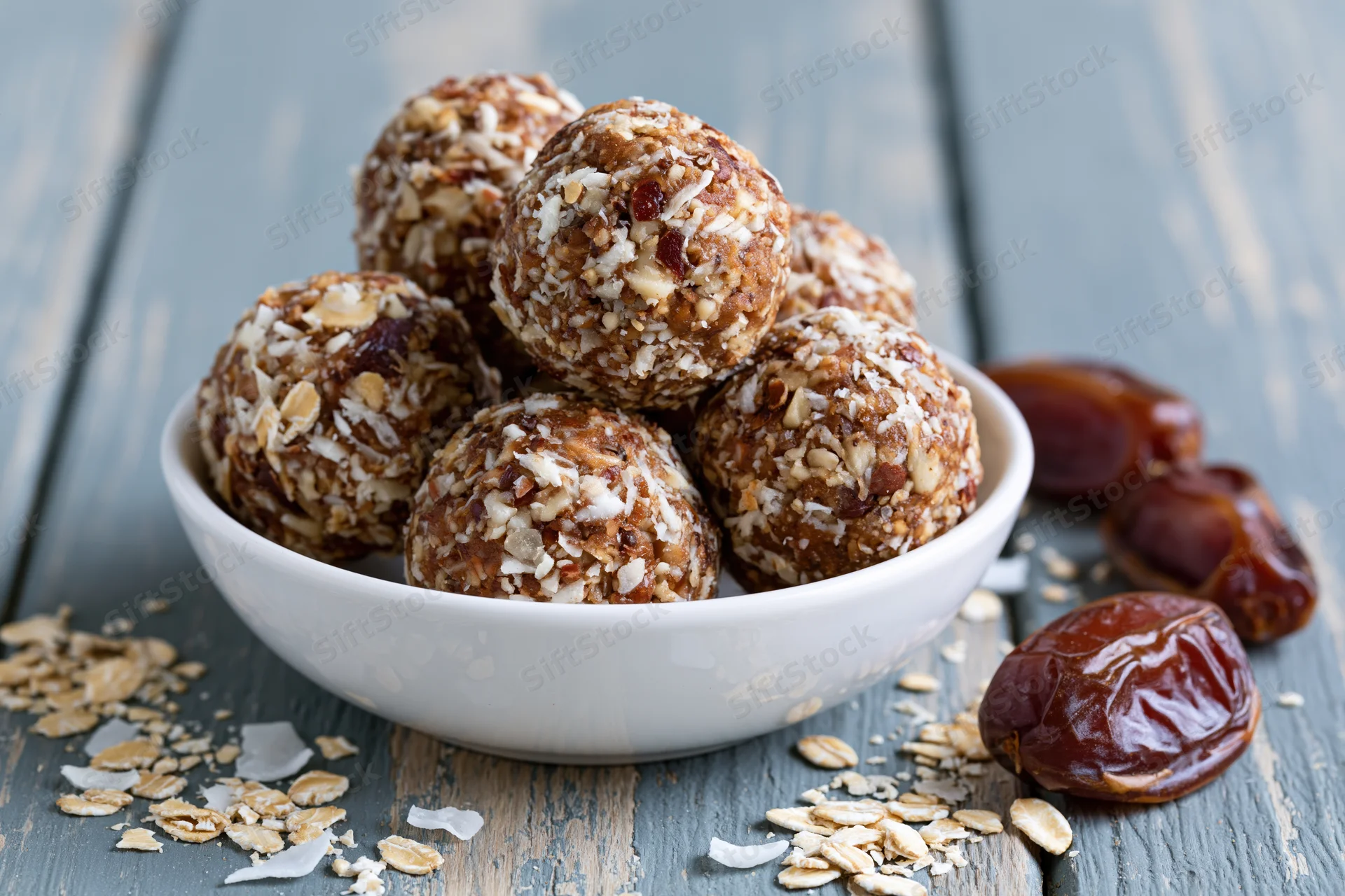 No-bake oat and date energy balls piled in a shallow white bowl on a rustic wooden table with scattered oats and dates nearby