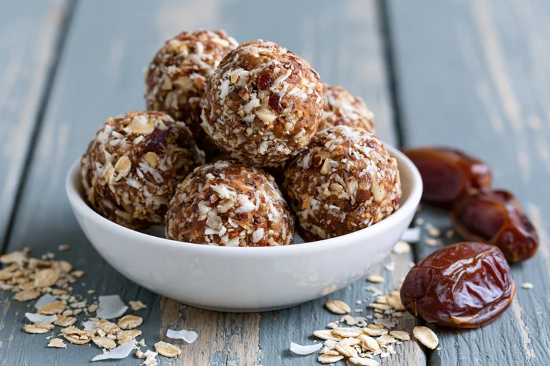 No-bake oat and date energy balls piled in a shallow white bowl on a rustic wooden table with scattered oats and dates nearby