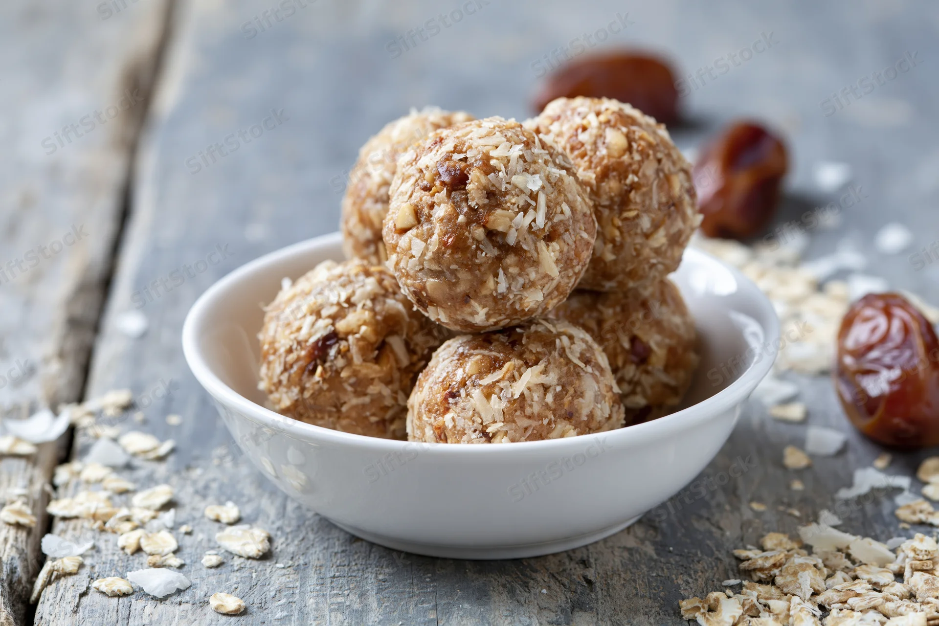 No-bake oat and date energy balls piled in a shallow white bowl on a rustic wooden surface with scattered oats and dates around