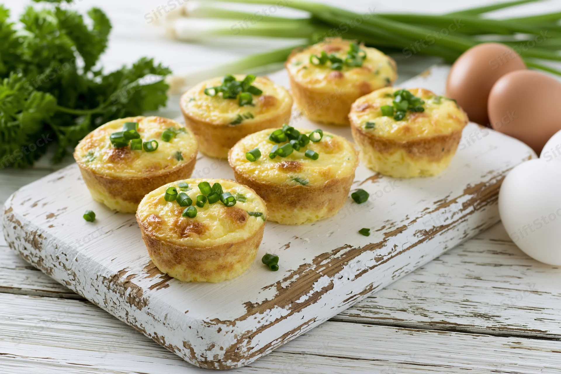 Six baked egg muffins topped with chopped green onions arranged on a white distressed wooden cutting board