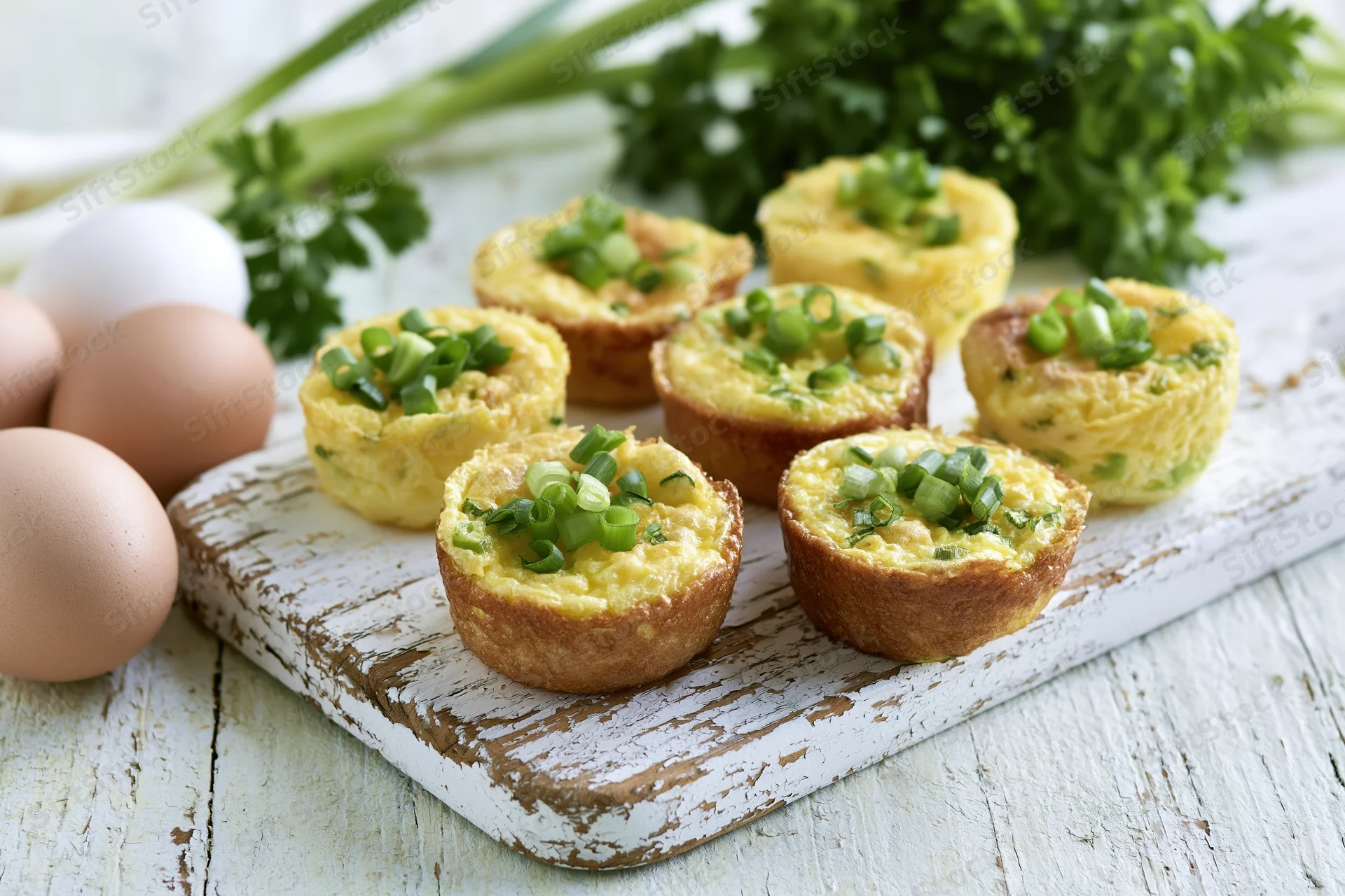 Baked egg muffins topped with chopped green onion arranged on a rustic white wooden cutting board with whole eggs and fresh herbs nearby
