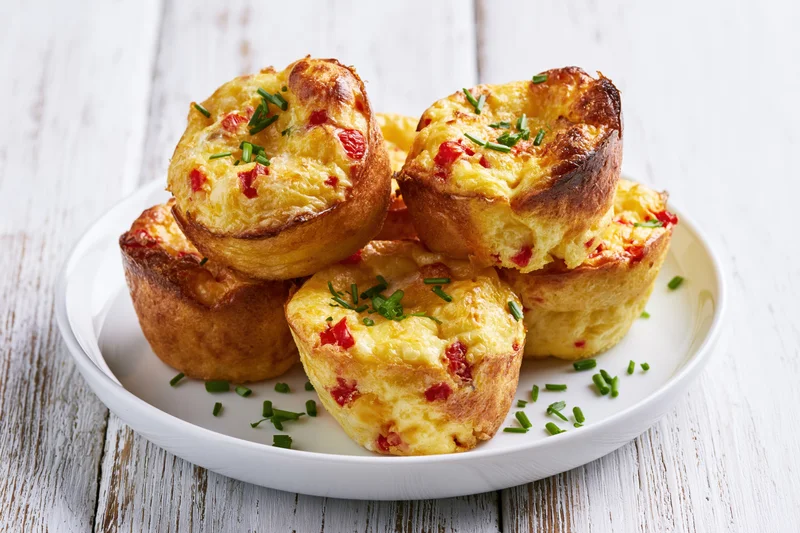 Golden-brown egg muffin bites stacked on a white plate with chives and red pepper on a wooden surface