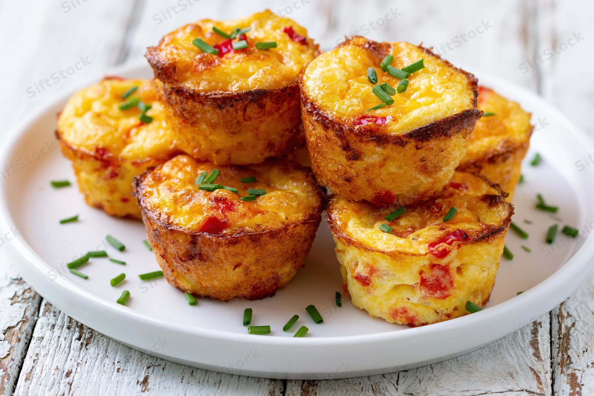 Golden-brown egg muffin bites stacked on a white plate with chives and bacon bits