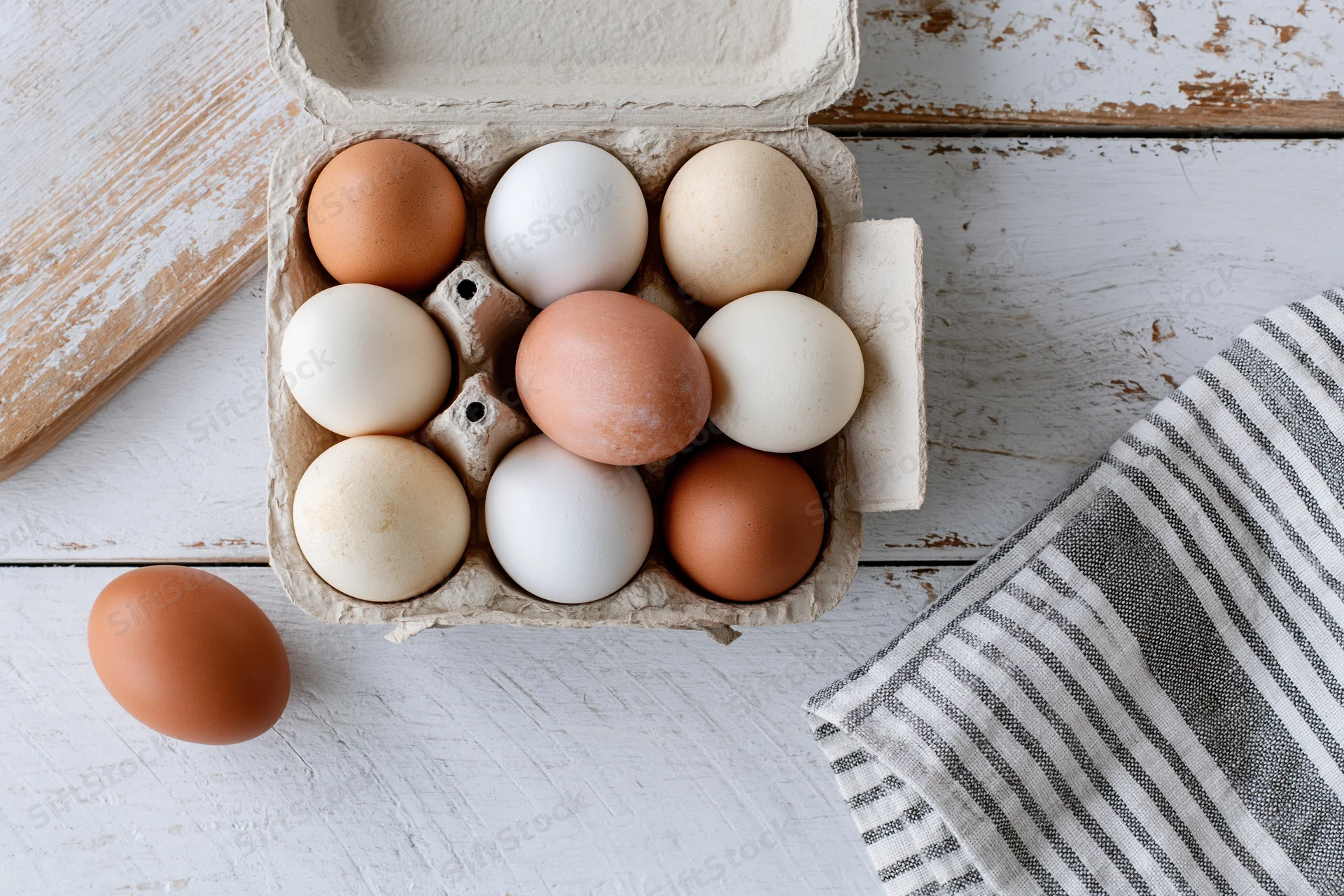Mixed brown and white eggs in open cardboard carton on white wooden table with striped towel