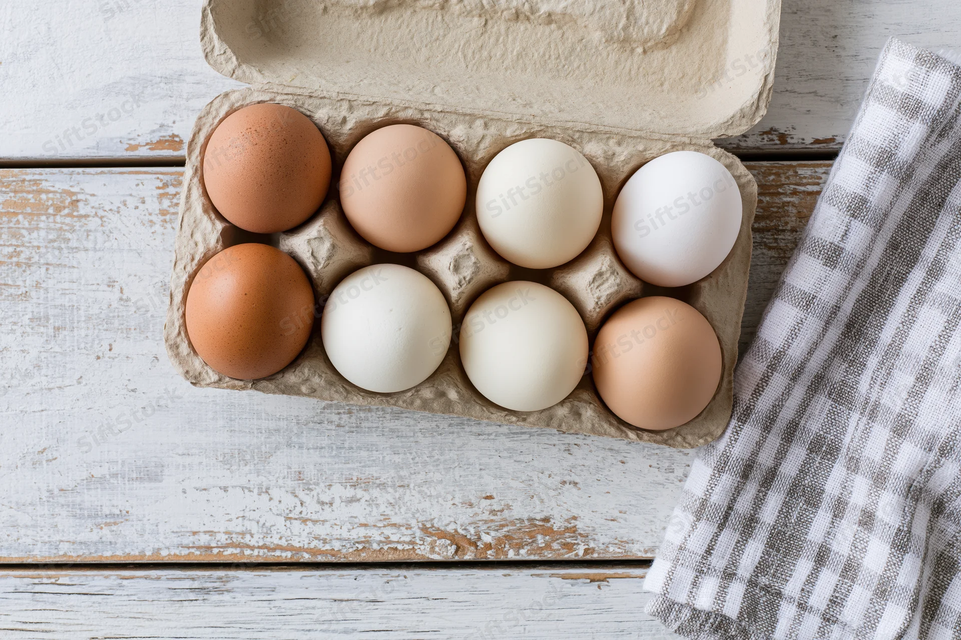 Six mixed brown and white eggs in an open cardboard carton on a rustic wooden table with a checkered cloth beside it