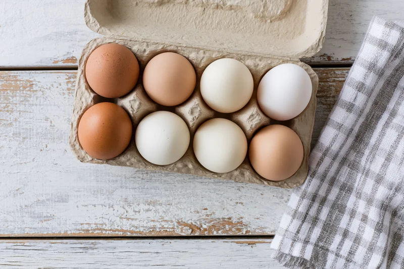 Six mixed brown and white eggs in an open cardboard carton on a rustic wooden table with a checkered cloth beside it
