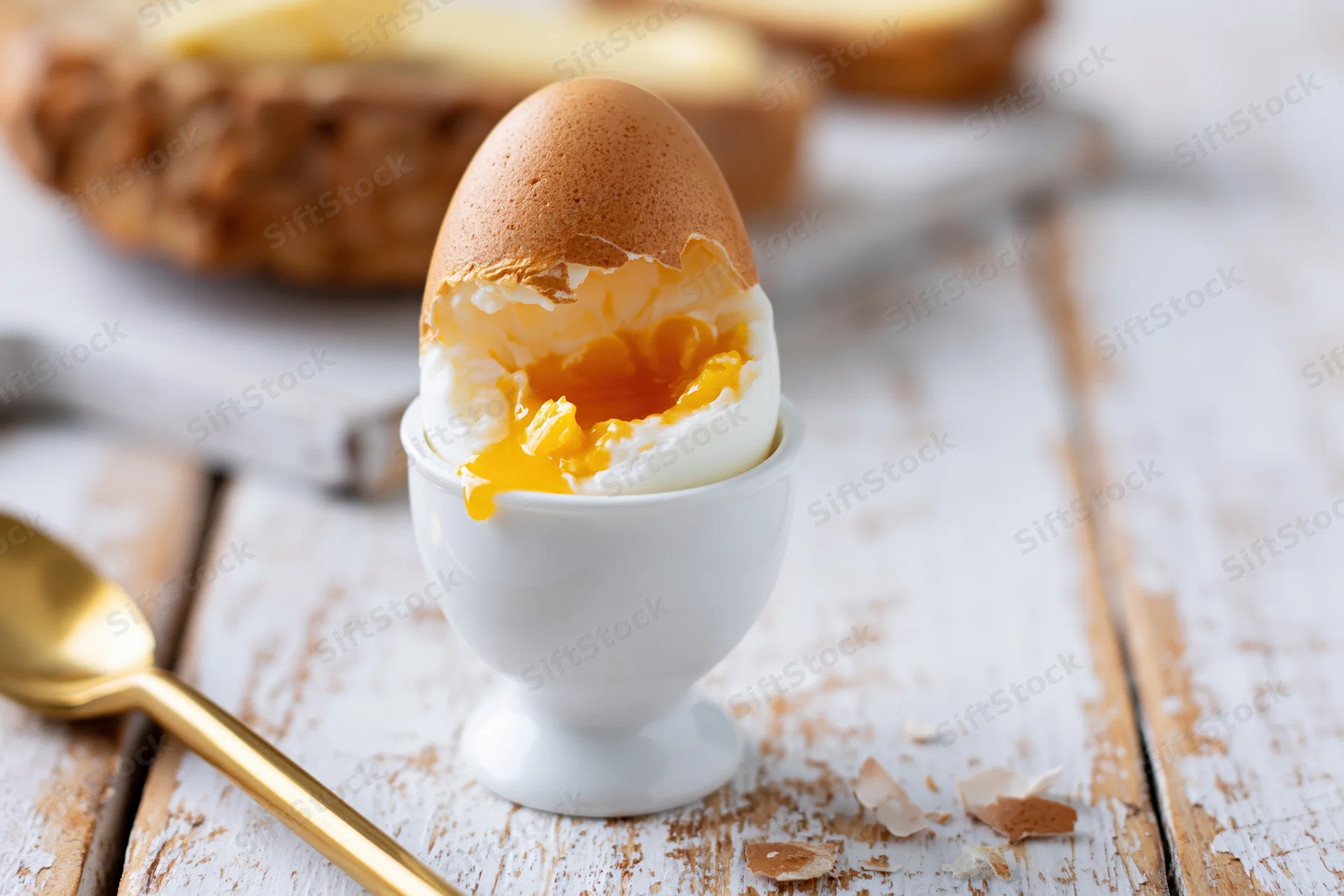 Soft-boiled egg cracked open in a white ceramic eggcup on a rustic wooden table with a golden spoon beside it