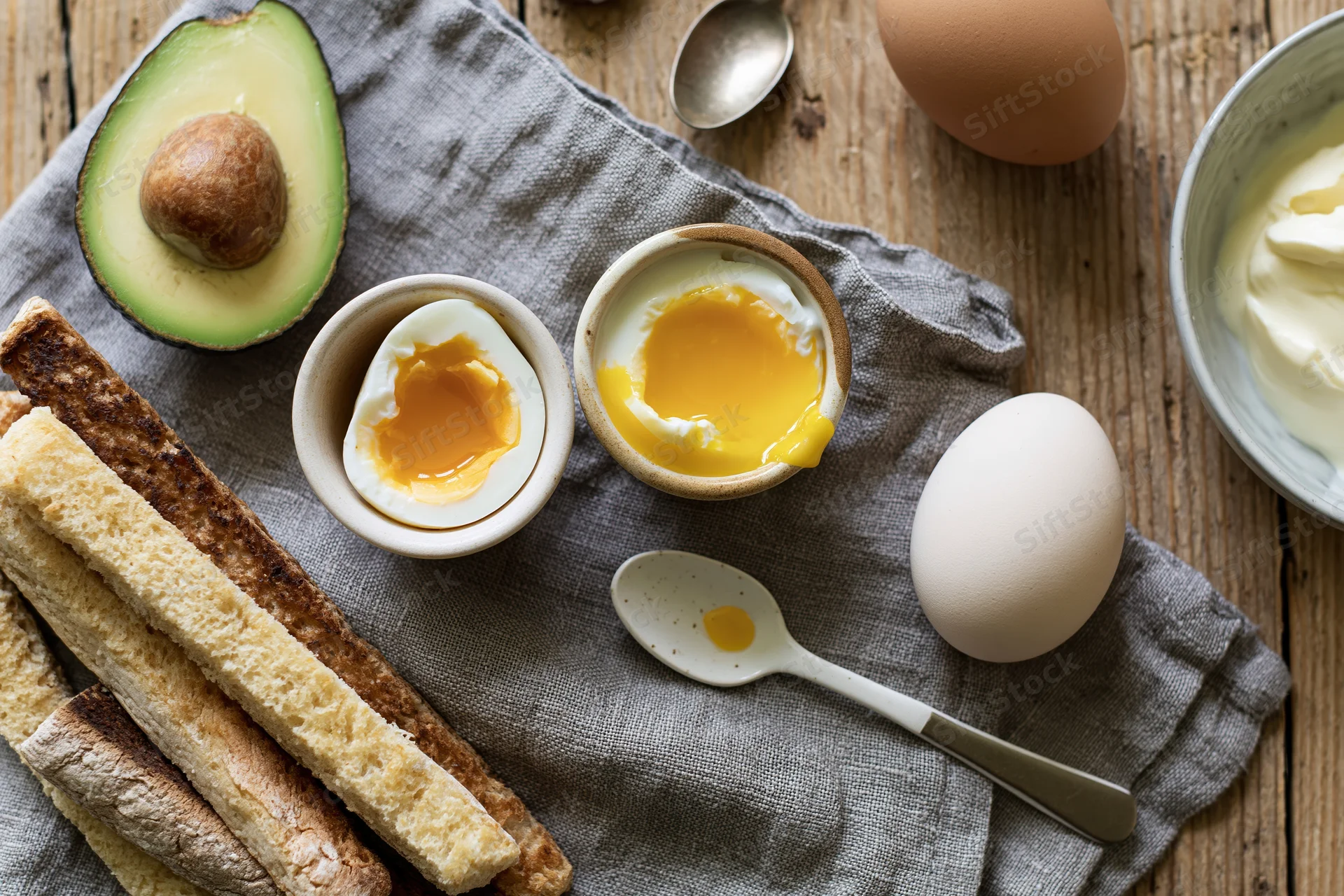 Soft-boiled egg cracked open in a ceramic egg cup with runny yolk on a wooden table with avocado, toast, and yogurt nearby