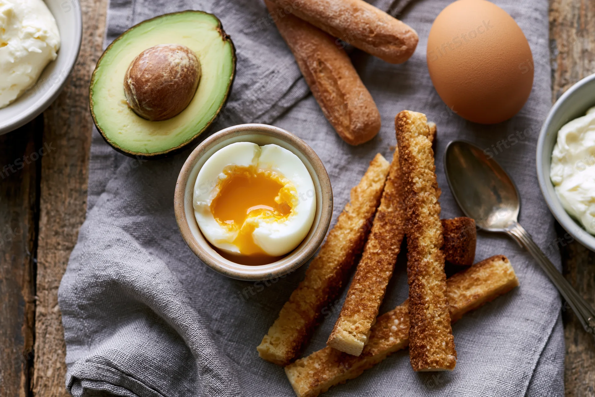 Soft-boiled egg cracked open in a ceramic egg cup with runny yolk, surrounded by avocado half, toast sticks, cream, and spoon on a gray cloth over wooden surface