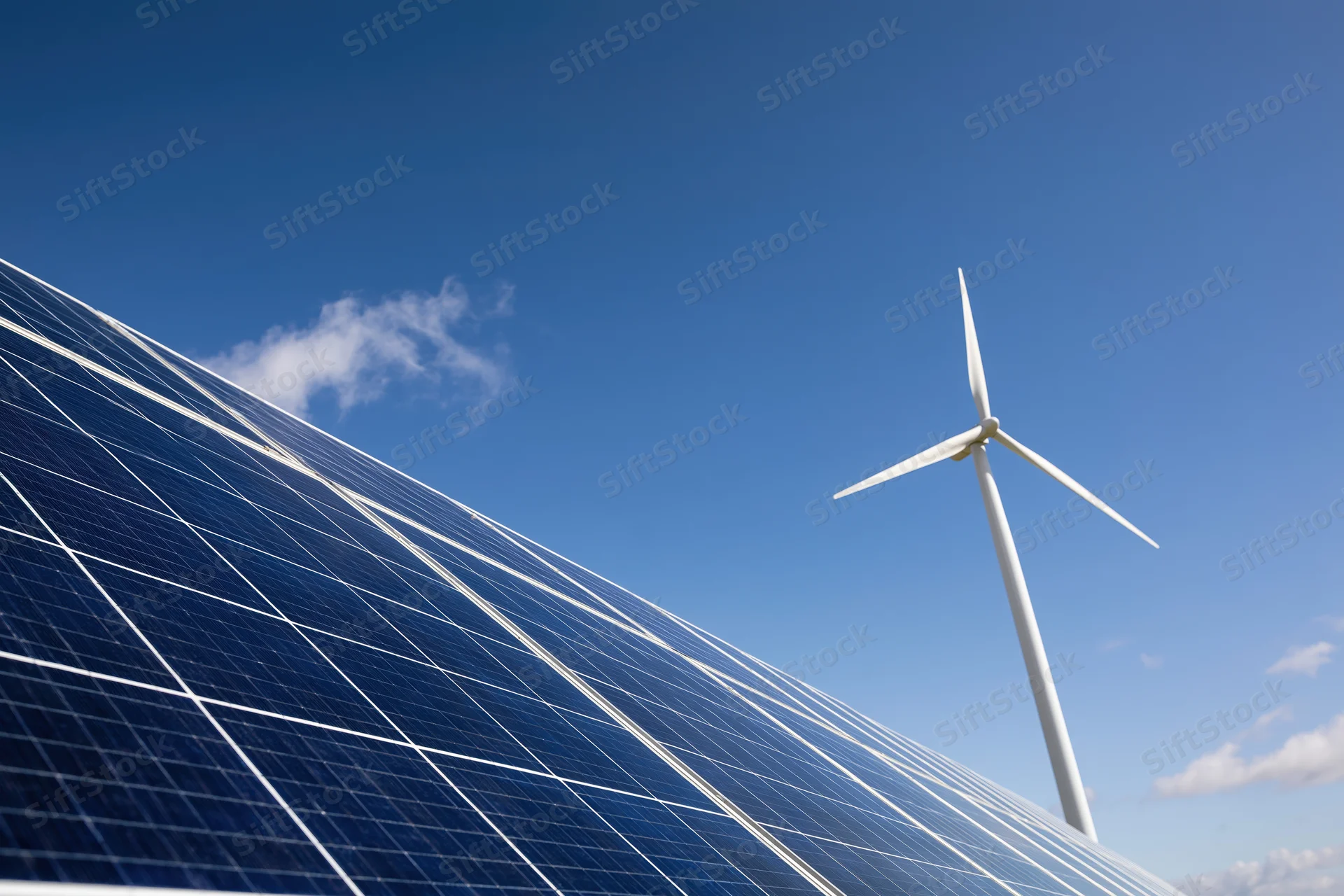 Solar panel array filling the lower-left foreground with a wind turbine in the background under a clear blue sky