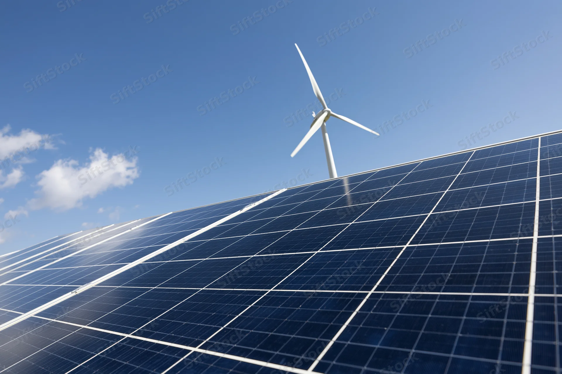 Solar panel array filling the foreground with a wind turbine in the background under a clear blue sky