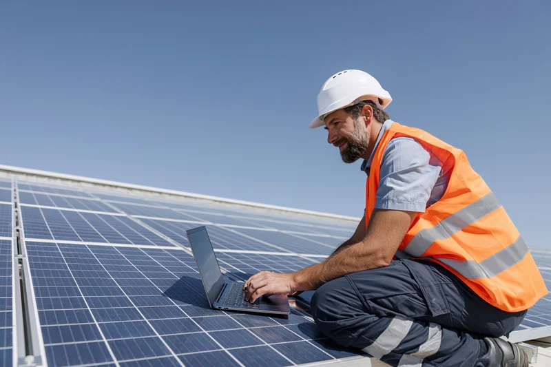 Solar technician crouching on rooftop solar array using laptop for inspection
