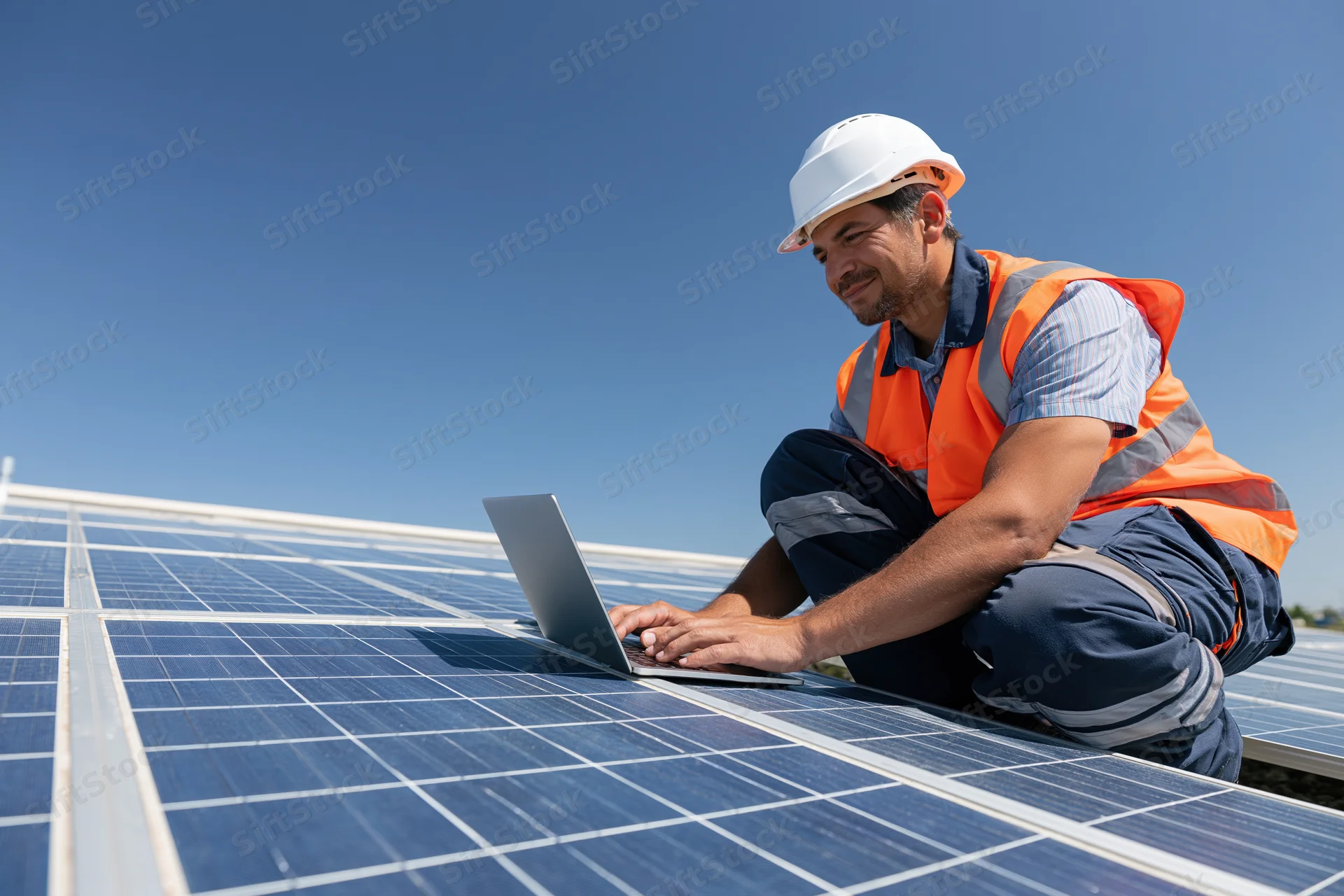 Technician crouching on rooftop solar array using laptop for inspection