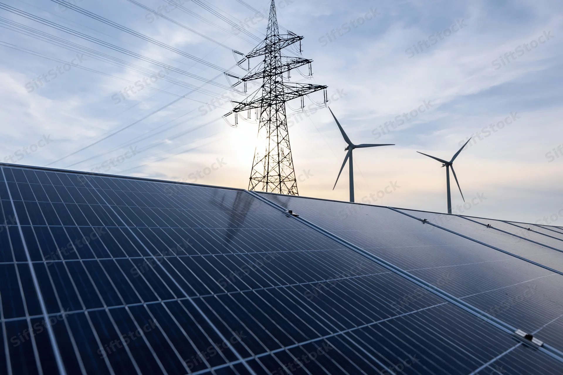 Solar panels in foreground with wind turbines and power transmission tower in background at sunset