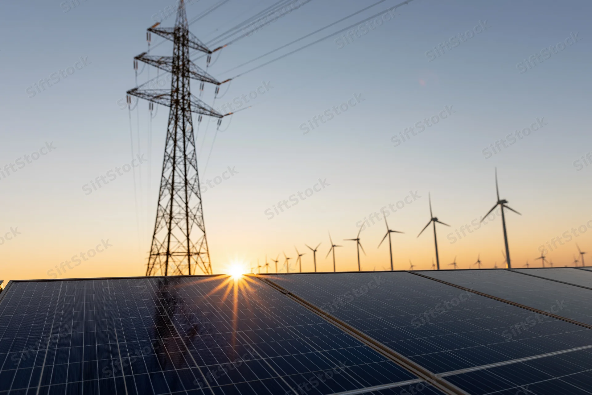 Solar panels reflecting golden sunset light with wind turbines and power transmission tower in background