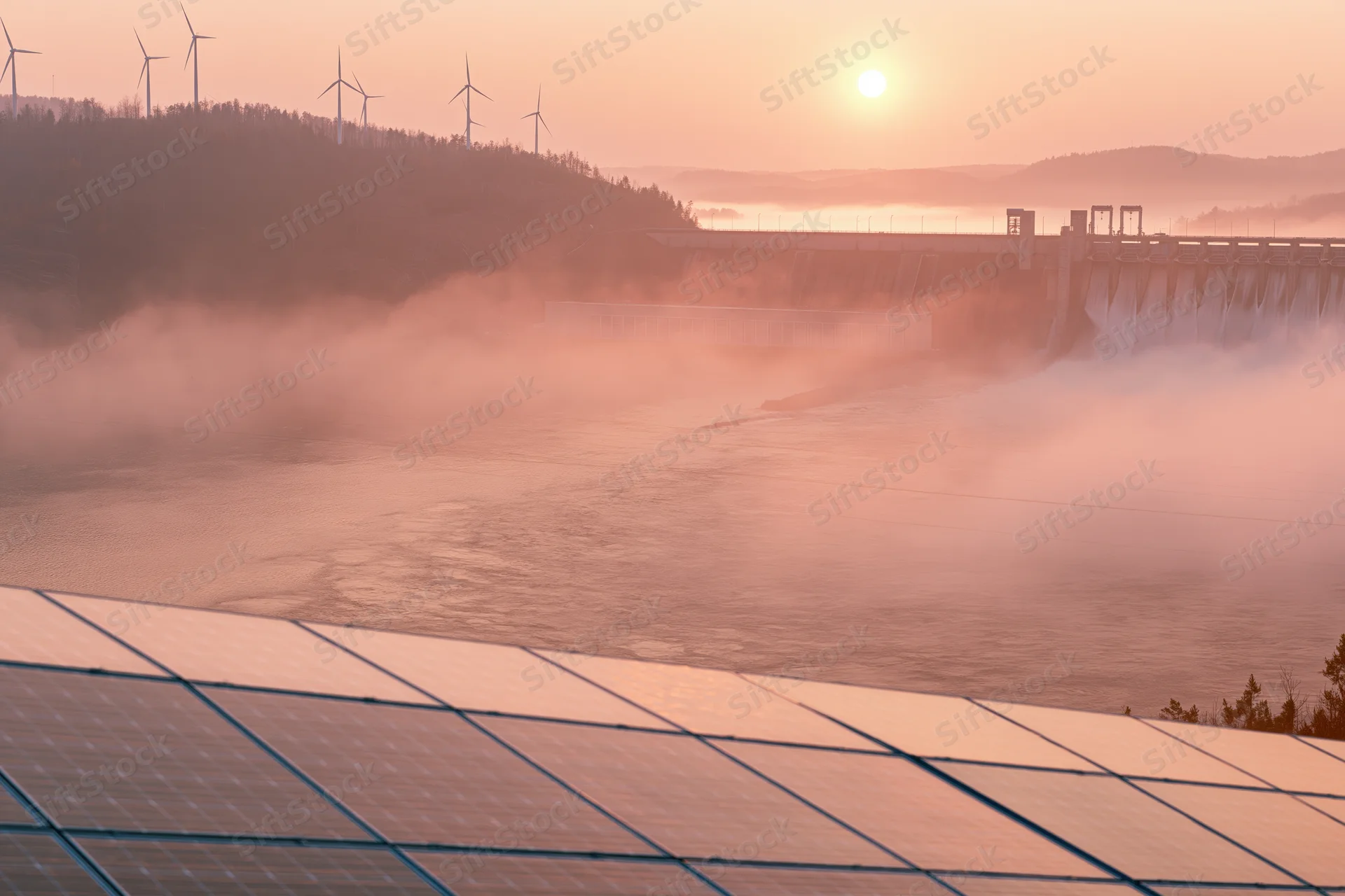 Solar panels in foreground with hydroelectric dam and wind turbines on hillside at sunrise