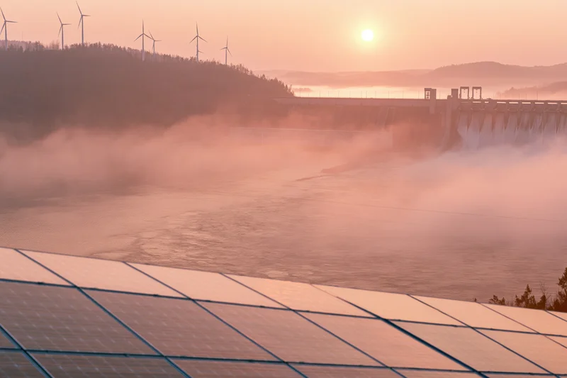 Solar panels in foreground with hydroelectric dam and wind turbines on hillside at sunrise