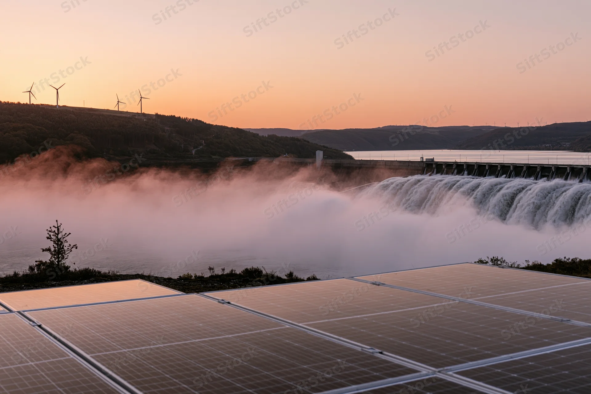 Solar panels in foreground with hydroelectric dam and waterfall at sunset, wind turbines on hills in distance