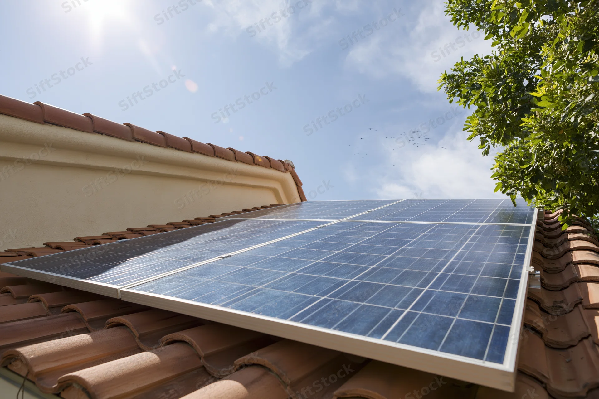 Solar panels installed on a terracotta-tiled residential roof under clear blue sky with tree foliage visible