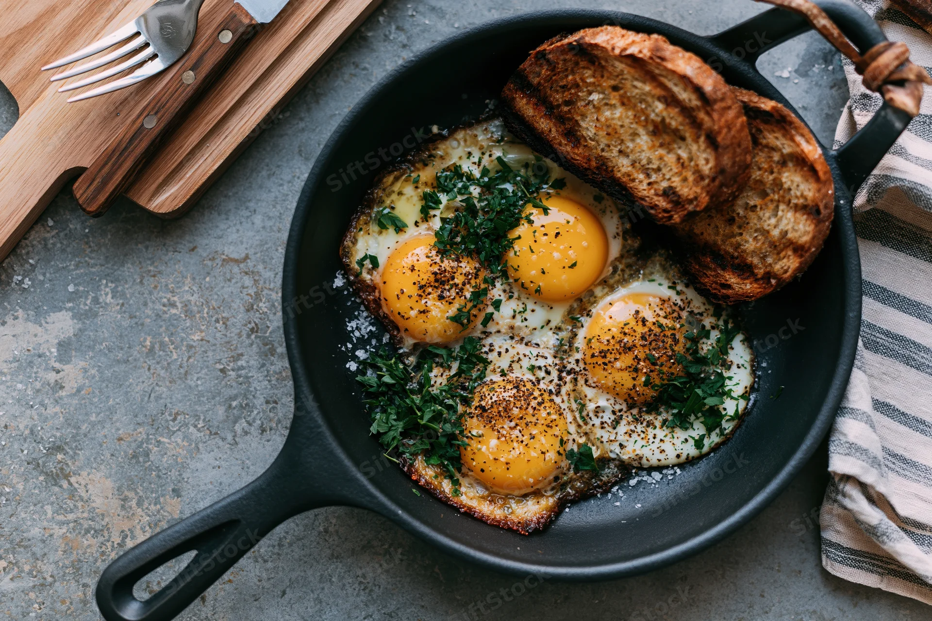 Three fried eggs with golden yolks in a cast iron skillet with toast and herbs on a gray countertop
