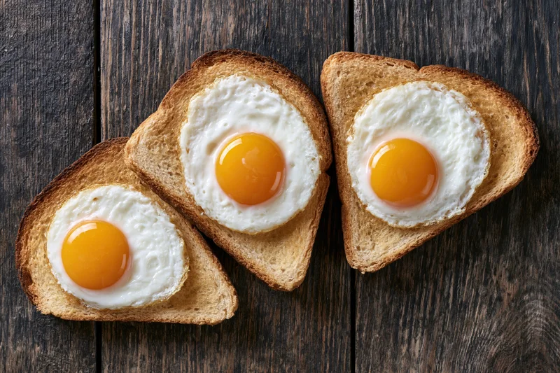 Three slices of toast each topped with a sunny-side-up fried egg on a dark wooden surface
