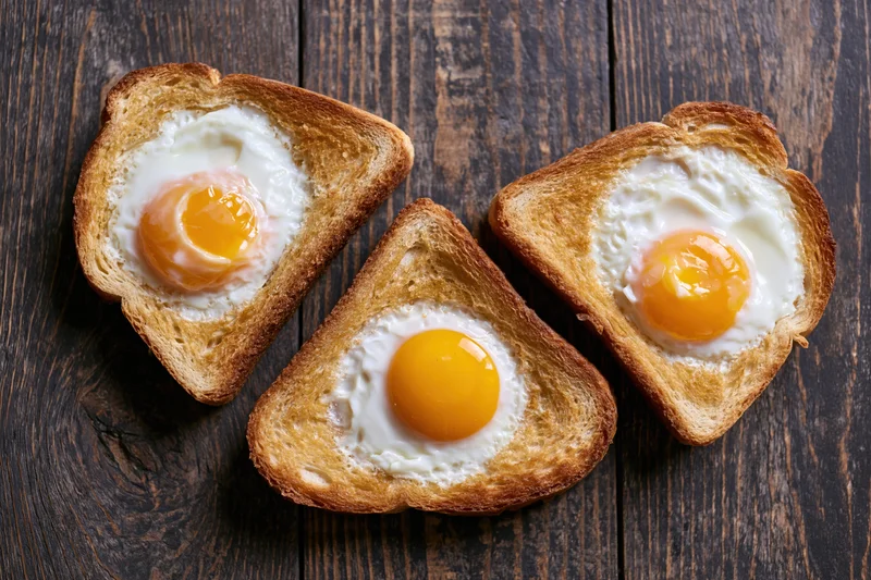 Three toast slices with fried eggs cooked in center holes on dark wooden surface