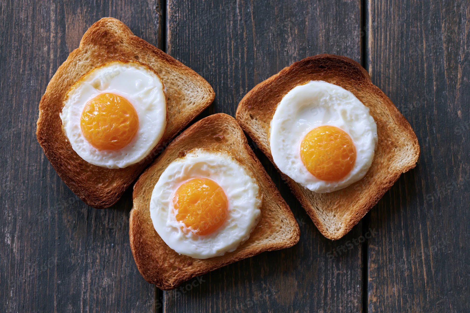 Three slices of toast each topped with a fried egg arranged on a dark wooden surface