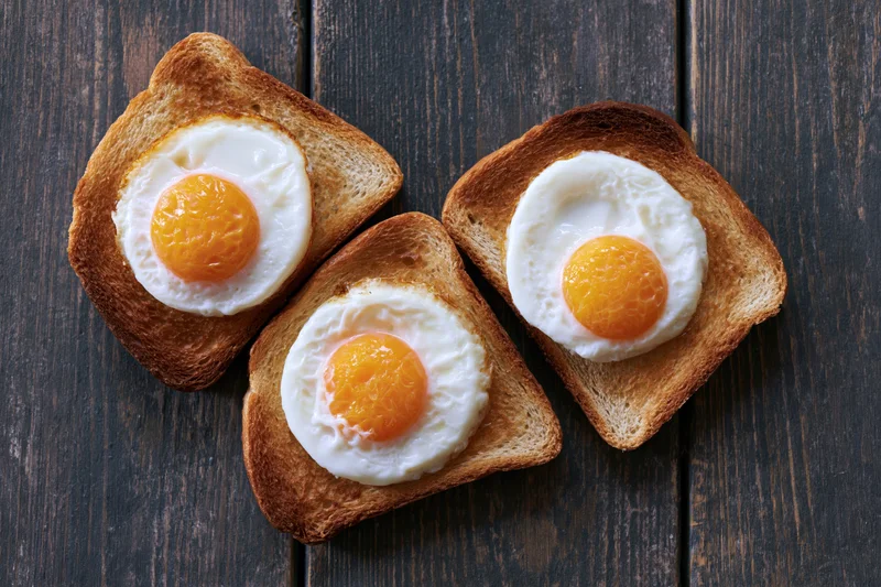 Three slices of toast each topped with a fried egg arranged on a dark wooden surface