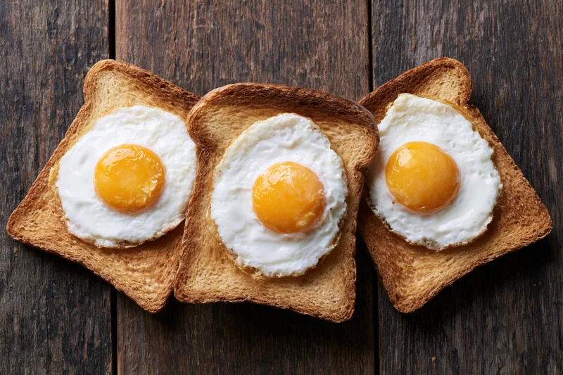 Three slices of toast each topped with a fried egg arranged on a dark wooden surface
