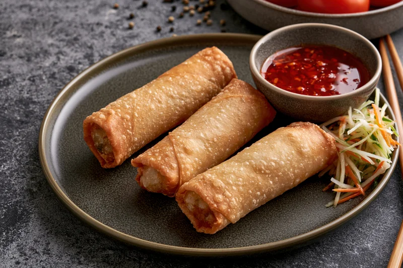 Three golden fried spring rolls arranged on a dark olive ceramic plate with dipping sauce and vegetable garnish