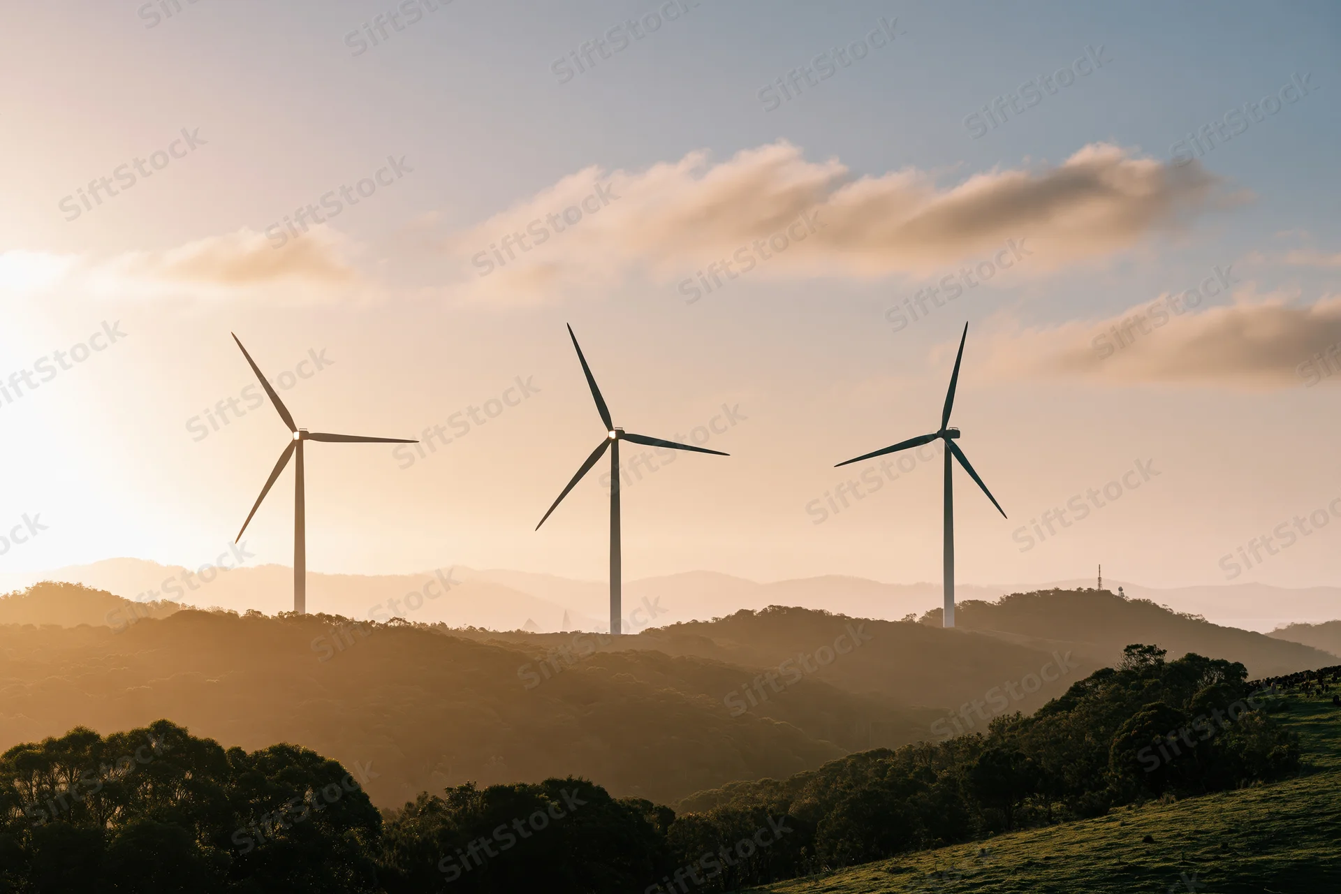 Three wind turbines silhouetted against a golden sky above rolling forested hills at sunset