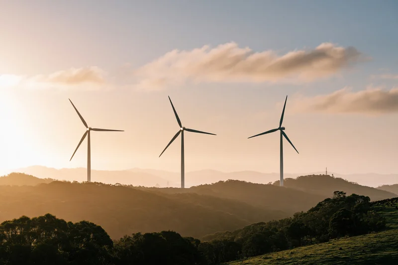 Three wind turbines silhouetted against a golden sky above rolling forested hills at sunset