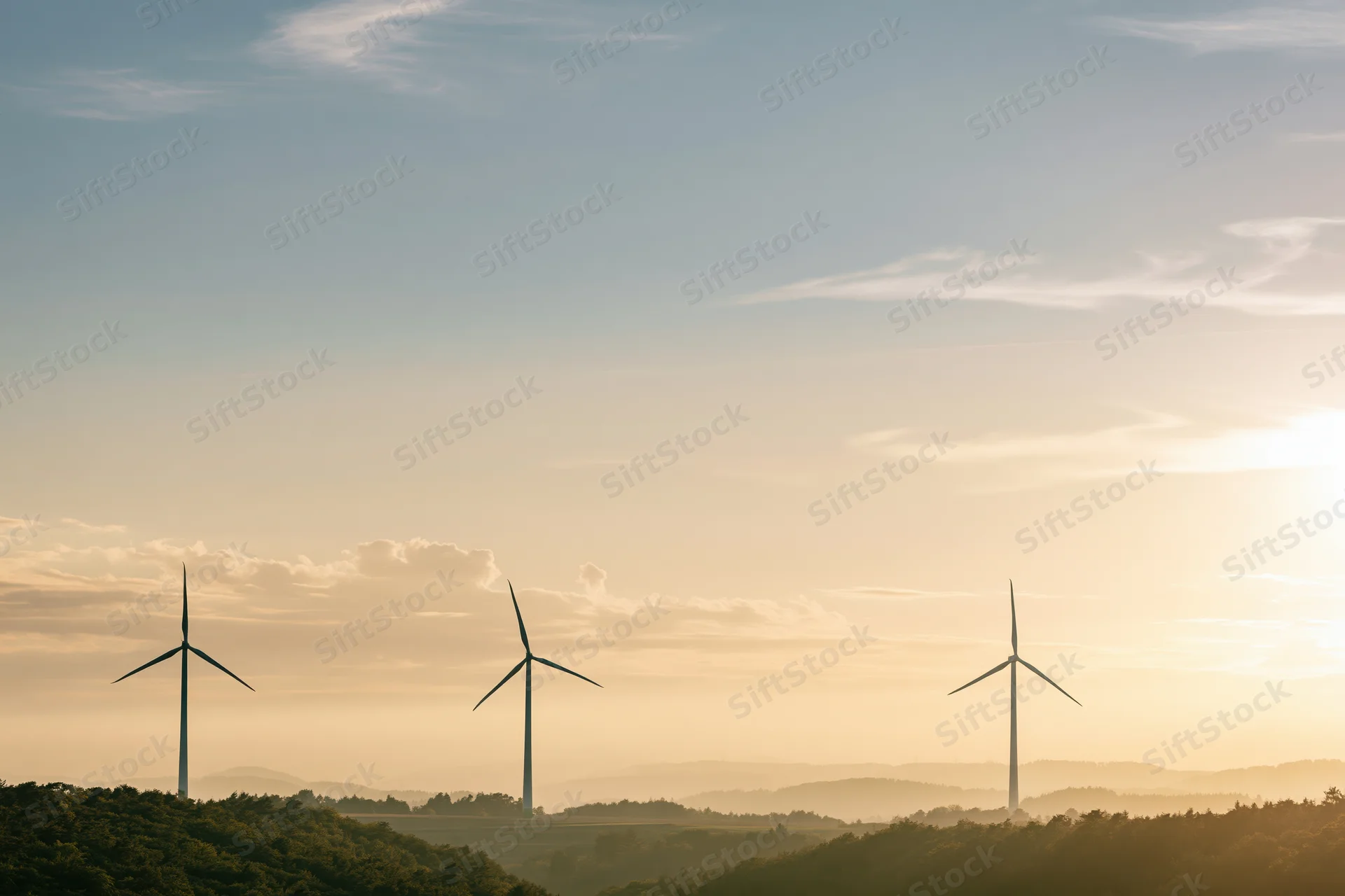Three wind turbines silhouetted against a golden sky above rolling forested hills