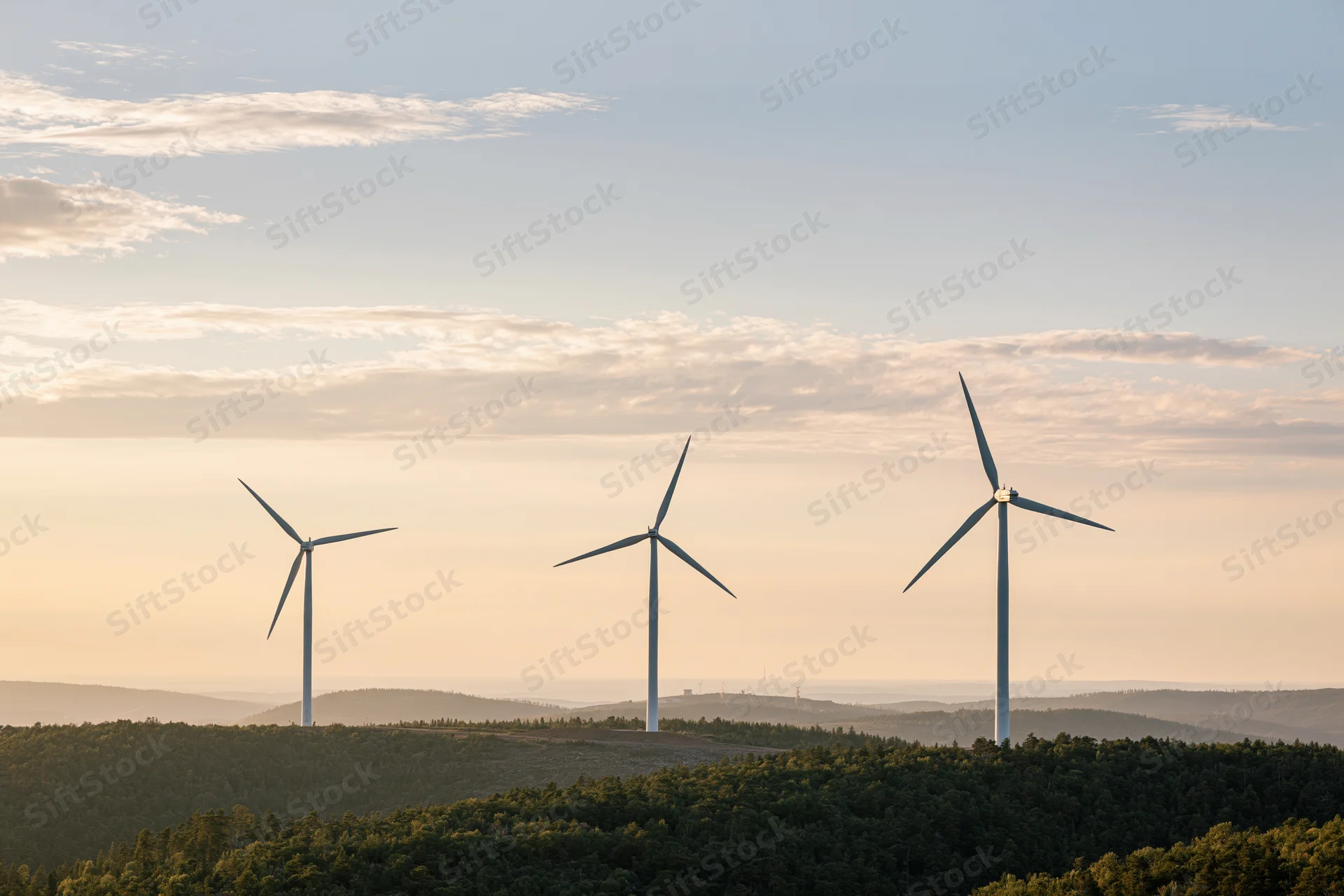Three wind turbines rising above rolling green forested hills at sunset