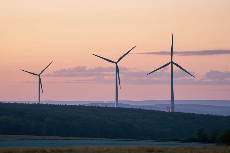 Three wind turbines standing on a hillside at sunset over a forested valley