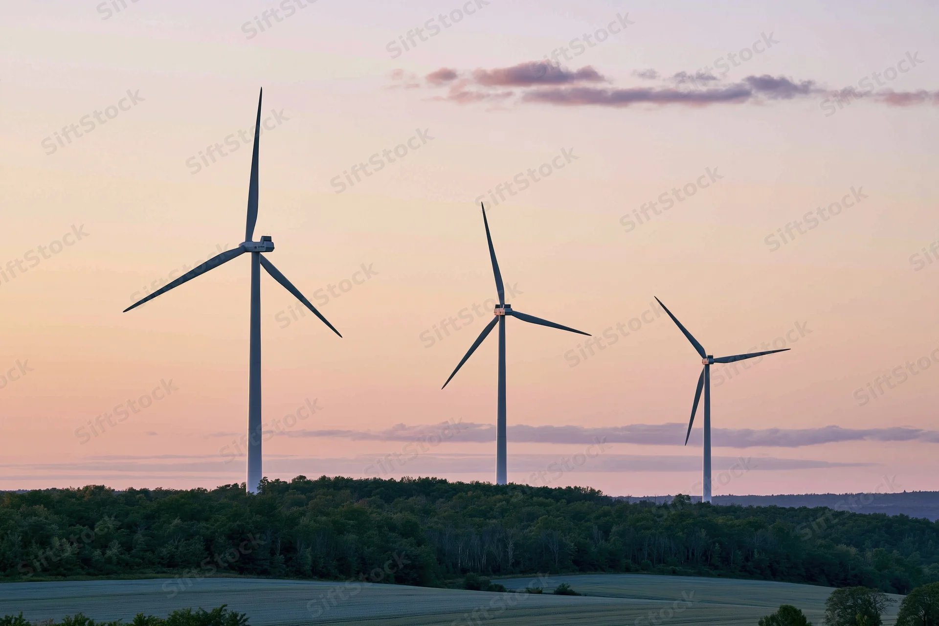 Three wind turbines standing in a row on a tree-lined hillside at sunset
