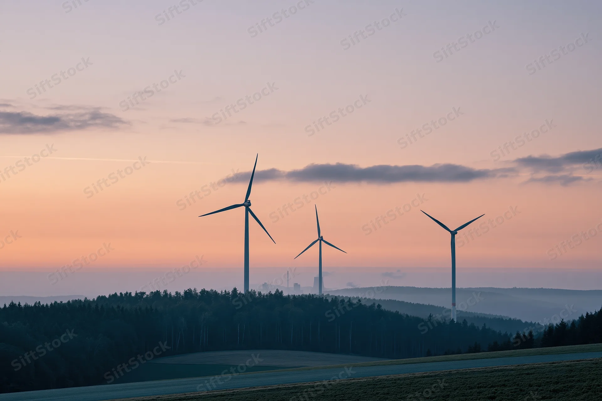Three wind turbines standing on a rolling hillside at sunset with forest and fields below