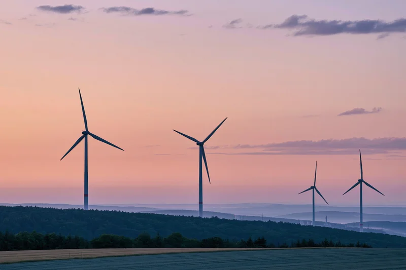Wind turbines standing on a rolling hillside at sunset with fields and forest below